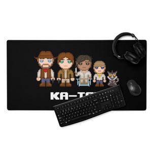 May include: A black gaming mouse pad with a pixelated image of five characters and a small animal. The text "KA-T" is printed below the characters. A black keyboard and a pair of black headphones are also in the image.