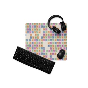 Può includere: Una tastiera nera, un mouse e un paio di cuffie over-ear sono su un tappetino per mouse con un design di tavola periodica. I quadrati della tavola periodica sono di vari colori, tra cui rosa, blu e giallo.