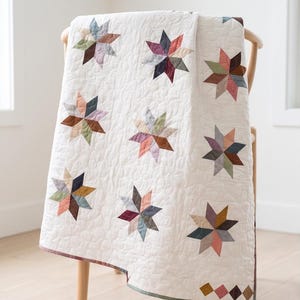 May include: A white quilt featuring a repeating pattern of multi-colored eight-pointed stars in shades of muted pink, blue, brown, and mustard yellow. The fabric has a textured, wavy quilted stitch pattern throughout and a thin border at the bottom.