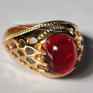 May include: A gold ring with a red gemstone set in a decorative, openwork design. The ring has a textured band and a round, cabochon-cut stone.