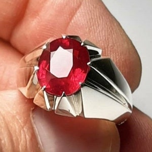 Op de afbeelding: Een zilveren ring met een grote, ovale, rode edelsteen in het midden. De ring heeft een gedetailleerd, geometrisch ontwerp.