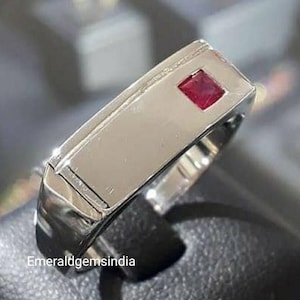 Op de afbeelding: Een gepolijste zilverkleurige ring met een plat, rechthoekig bovenoppervlak en een enkele, kleine, vierkant geslepen rode edelsteen aan één kant. De ring heeft een gladde, metalen afwerking en de tekst Emeraldgemsindia is zichtbaar.