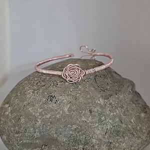 May include: A rose gold-coloured wire bracelet featuring a central rose-shaped wire design. The band is constructed from coiled wire and includes a hook-and-eye style closure mechanism. The metal has a metallic finish and a continuous circular shape.
