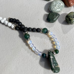 May include: A handmade necklace featuring a pendant and beads in various colors. The necklace includes white, black, green, and opalescent beads, with silver-colored metal accents. Several polished stones are scattered around the necklace.