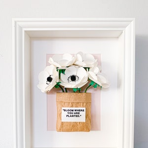 Op de afbeelding: Witte papieren bloemen in een bruine papieren zak met de tekst "BLOOM WHERE YOU ARE PLANTED." ingelijst in een wit frame.