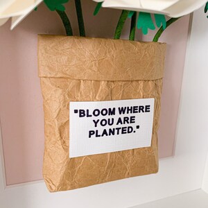 Op de afbeelding: Witte papieren bloemen in een bruine papieren zak met de tekst "Bloom where you are planted."