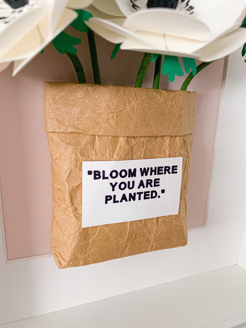 Op de afbeelding: Witte papieren bloemen in een bruine papieren zak met de tekst "Bloom where you are planted."
