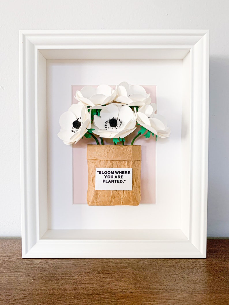 Op de afbeelding: Witte papieren bloemen in een bruine papieren zak met de tekst "BLOOM WHERE YOU ARE PLANTED." ingelijst in een wit frame.