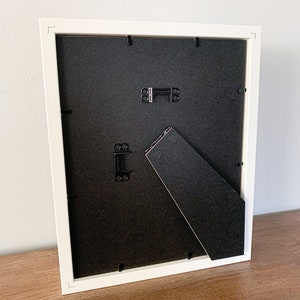 Op de afbeelding: Wit fotolijstje met een zwarte passe-partout en een ezelstandaard aan de achterkant. Het lijstje staat op een houten oppervlak.