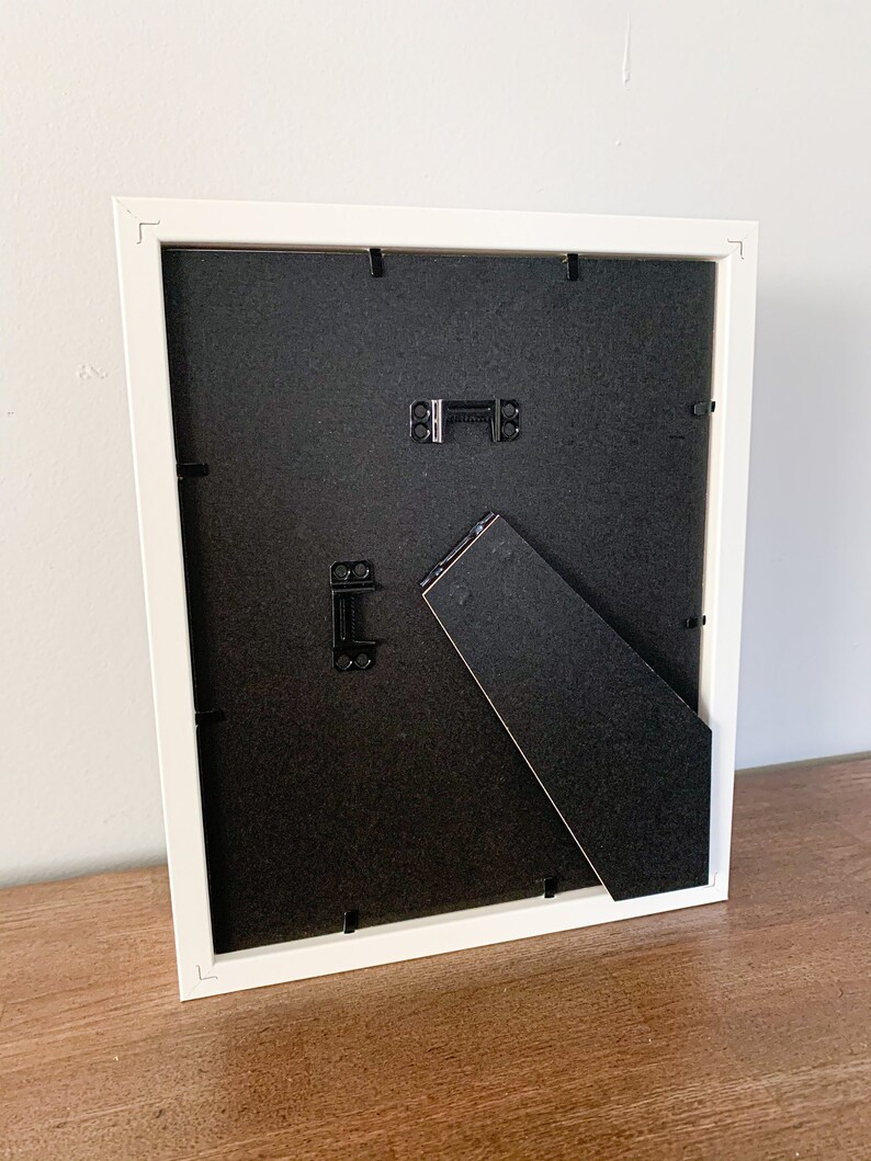 Op de afbeelding: Wit fotolijstje met een zwarte passe-partout en een ezelstandaard aan de achterkant. Het lijstje staat op een houten oppervlak.