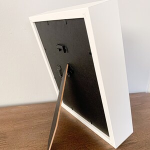 Op de afbeelding: Wit fotolijstje met een zwarte passe-partout en een houten ezelstandaard. Het lijstje staat op een houten oppervlak.