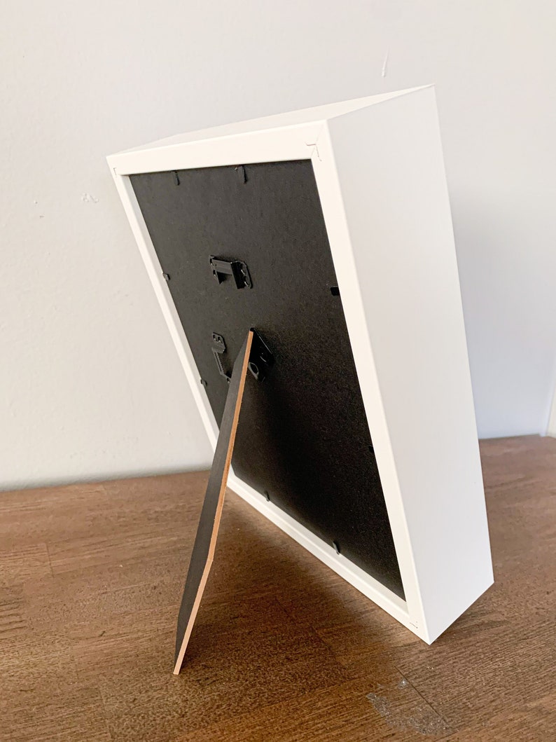 Op de afbeelding: Wit fotolijstje met een zwarte passe-partout en een houten ezelstandaard. Het lijstje staat op een houten oppervlak.