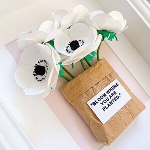 Op de afbeelding: Witte papieren bloemen in een bruine papieren zak met de quote "Bloom where you are planted." ingelijst in een wit frame.