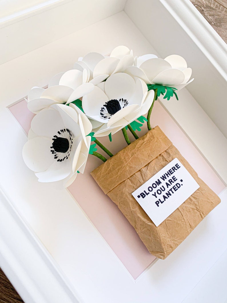 Op de afbeelding: Witte papieren bloemen in een bruine papieren zak met de quote "Bloom where you are planted." ingelijst in een wit frame.