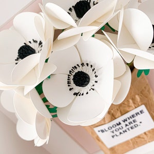 Op de afbeelding: Witte papieren bloemen met zwarte centra in een bruine papieren zak met de tekst "Bloom where you are planted."