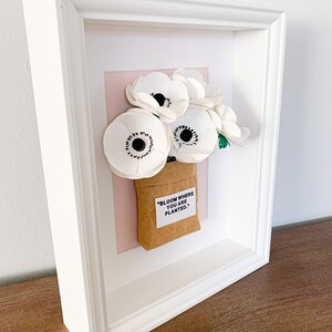 Op de afbeelding: Witte papieren bloemen in een bruine papieren zak met de quote "Bloom where you are planted." ingelijst in een wit frame met een roze achtergrond.
