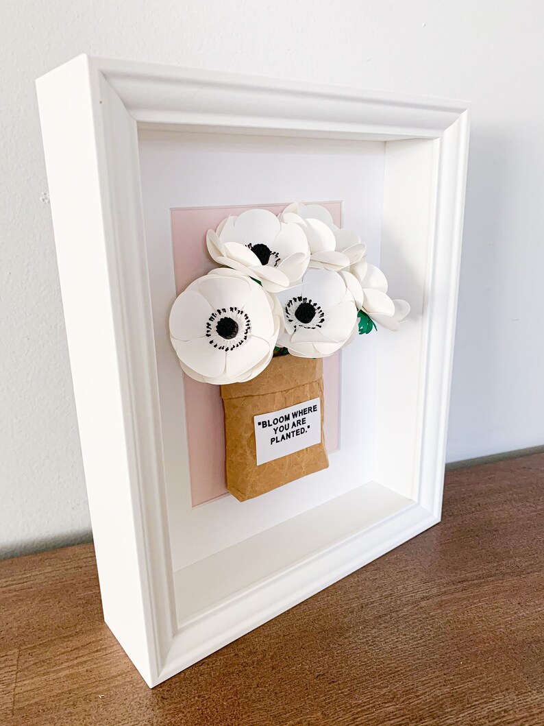 Op de afbeelding: Witte papieren bloemen in een bruine papieren zak met de quote "Bloom where you are planted." ingelijst in een wit frame met een roze achtergrond.