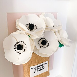 Op de afbeelding: Een ingelijst papieren bloemenarrangement in een bruine papieren zak met de quote "Bloom where you are planted." De bloemen zijn wit met zwarte centra en zijn gerangschikt op een roze achtergrond.
