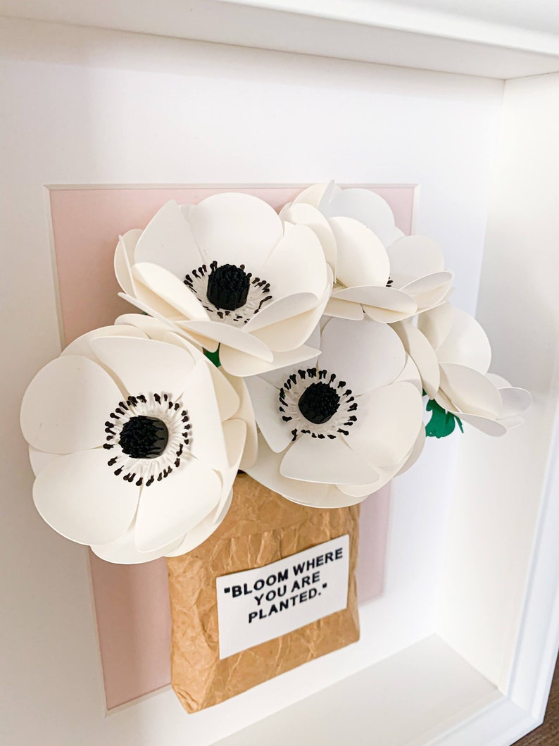 Op de afbeelding: Een ingelijst papieren bloemenarrangement in een bruine papieren zak met de quote "Bloom where you are planted." De bloemen zijn wit met zwarte centra en zijn gerangschikt op een roze achtergrond.