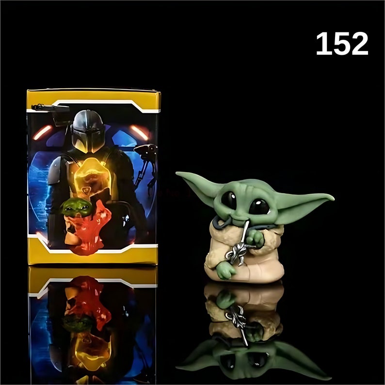 May include: A green and brown figurine of a character from the Star Wars franchise, known as "The Child" or "Baby Yoda". The figurine is holding a small, silver object in its hand. The figurine is sitting on a black surface. The image also shows a box with a picture of a character from the Star Wars franchise, known as "The Mandalorian".