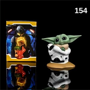 May include: A green and white figurine of a character from the Star Wars franchise, known as "The Child" or "Baby Yoda", holding a small white droid. The figurine is on a brown base. The image also shows a box with a picture of a Mandalorian character.