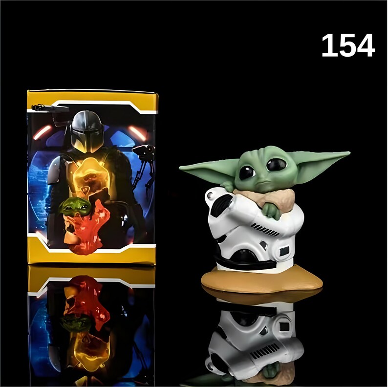May include: A green and white figurine of a character from the Star Wars franchise, known as "The Child" or "Baby Yoda", holding a small white droid. The figurine is on a brown base. The image also shows a box with a picture of a Mandalorian character.