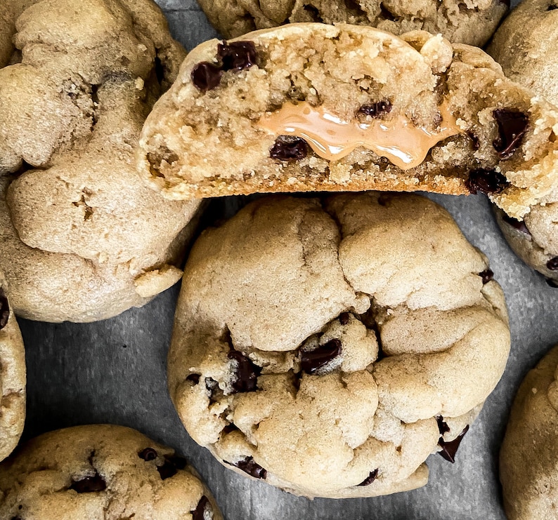 Puede incluir: Primer plano de varias galletas con chispas de chocolate reci&eacute;n horneadas. Una galleta est&aacute; cortada por la mitad, revelando un cremoso relleno de mantequilla de man&iacute;. Las galletas son de color marr&oacute;n dorado con chispas de chocolate oscuro, dispuestas sobre una superficie gris.