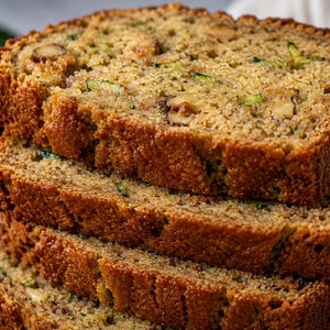May include: A stack of four thick slices of golden-brown zucchini bread featuring visible green flecks of zucchini and chopped brown walnuts throughout the crumb. The bread has a textured, porous surface and a slightly darker, baked crust edge.