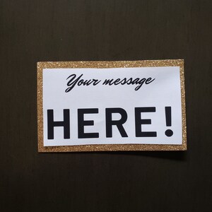 Puede incluir: Un letrero rectangular con un borde dorado brillante y un interior blanco. El texto "Your message" est&aacute; escrito en cursiva sobre el texto en negrita "HERE!" en negro. El letrero est&aacute; sobre un fondo marr&oacute;n oscuro.