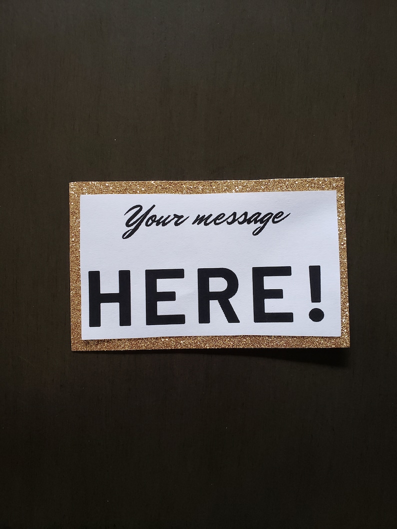 Puede incluir: Un letrero rectangular con un borde dorado brillante y un interior blanco. El texto "Your message" est&aacute; escrito en cursiva sobre el texto en negrita "HERE!" en negro. El letrero est&aacute; sobre un fondo marr&oacute;n oscuro.