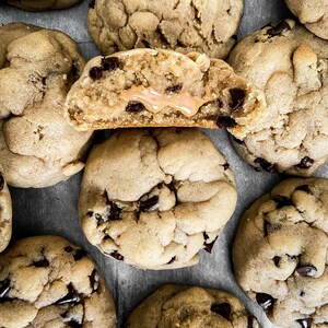 Puede incluir: Primer plano de varias galletas con chispas de chocolate, una cortada por la mitad para revelar un relleno de mantequilla de man&iacute;. Las galletas son de color marr&oacute;n dorado con chispas de chocolate oscuro.