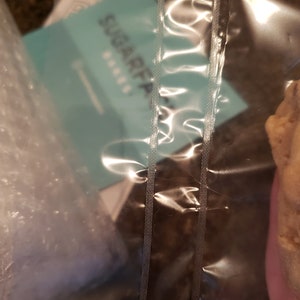 Puede incluir: Una bolsa de pl&aacute;stico transparente sellada con costuras verticales, que contiene una tarjeta turquesa con el texto "SUGARFAIR BAKES". La bolsa tambi&eacute;n contiene pl&aacute;stico de burbujas y un producto horneado, parcialmente visible a la derecha.