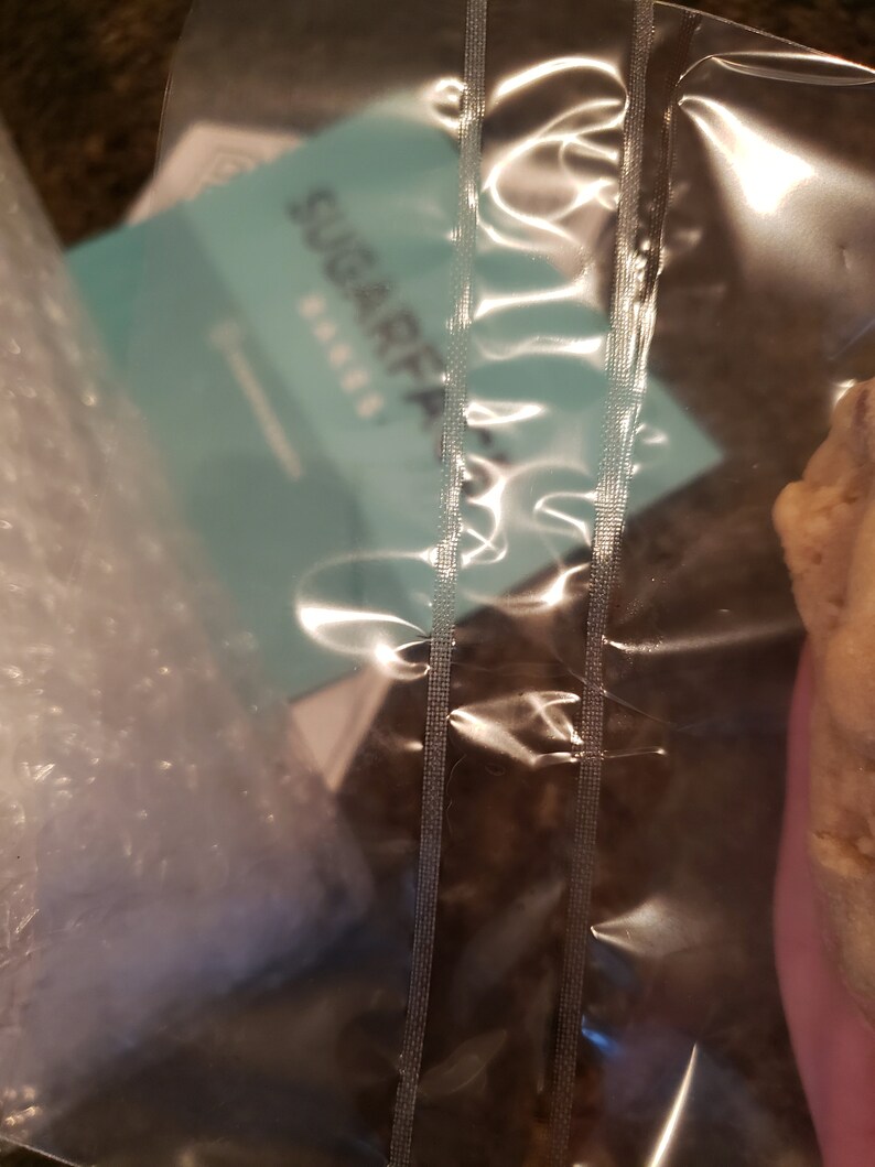 Puede incluir: Una bolsa de pl&aacute;stico transparente sellada con costuras verticales, que contiene una tarjeta turquesa con el texto "SUGARFAIR BAKES". La bolsa tambi&eacute;n contiene pl&aacute;stico de burbujas y un producto horneado, parcialmente visible a la derecha.