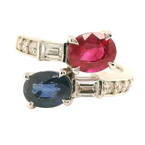 Może przedstawiać: Pierścionek z białego złota z czerwonym rubinem i niebieskim szafirem, oba osadzone w owalnych kształtach. Pierścionek jest również ozdobiony małymi diamentami.