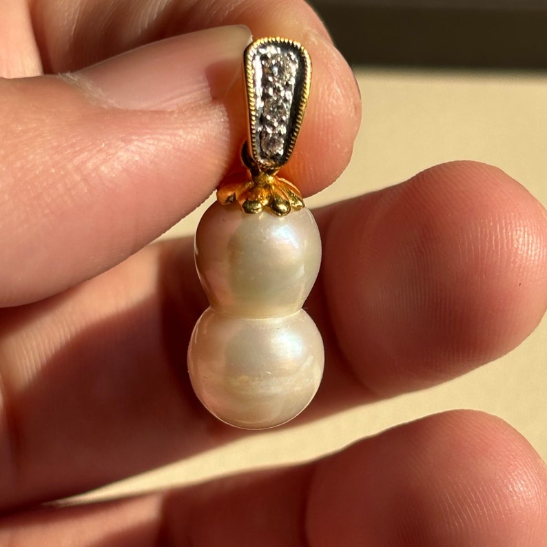 May include: A gold pendant featuring two white pearls, one larger than the other, with a small diamond-encrusted bail.