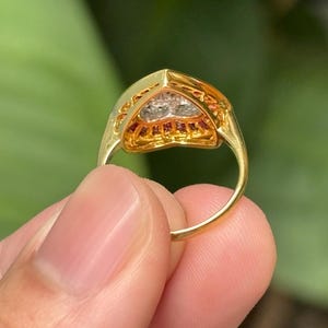 Pu&ograve; includere: Un anello d'oro con un'incastonatura triangolare. L'incastonatura &egrave; incrostata di piccole pietre preziose rosse e chiare. L'anello &egrave; tenuto contro uno sfondo verde sfocato.