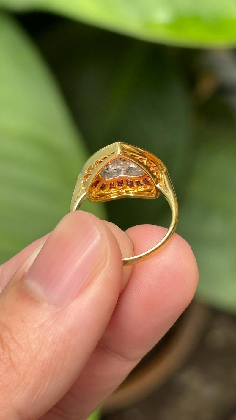 Pu&ograve; includere: Un anello d'oro con un'incastonatura triangolare. L'incastonatura &egrave; incrostata di piccole pietre preziose rosse e chiare. L'anello &egrave; tenuto contro uno sfondo verde sfocato.