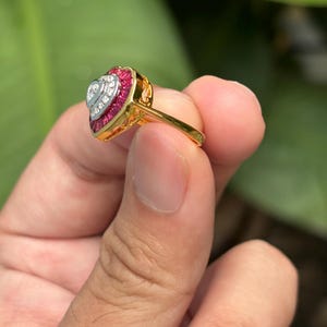 Pu&ograve; includere: Un anello d'oro con un design a forma di cuore. L'anello presenta un cuore centrale di pietre preziose bianche, circondato da un bordo di pietre preziose rosse. La fascia dell'anello &egrave; in oro lucido.