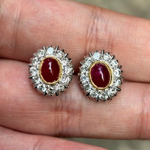 May include: A pair of ruby and diamond earrings. Each earring features a red oval-shaped ruby surrounded by a halo of small diamonds. The diamonds are set in a white metal, and the ruby is set in a yellow metal.