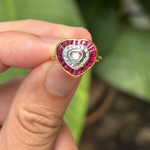 Pu&ograve; includere: Un anello a forma di cuore con una fascia d'oro. L'anello presenta un design centrale a forma di cuore con un diamante al centro, circondato da diamanti pi&ugrave; piccoli e pietre preziose rosse. L'anello &egrave; tenuto su uno sfondo verde sfocato.