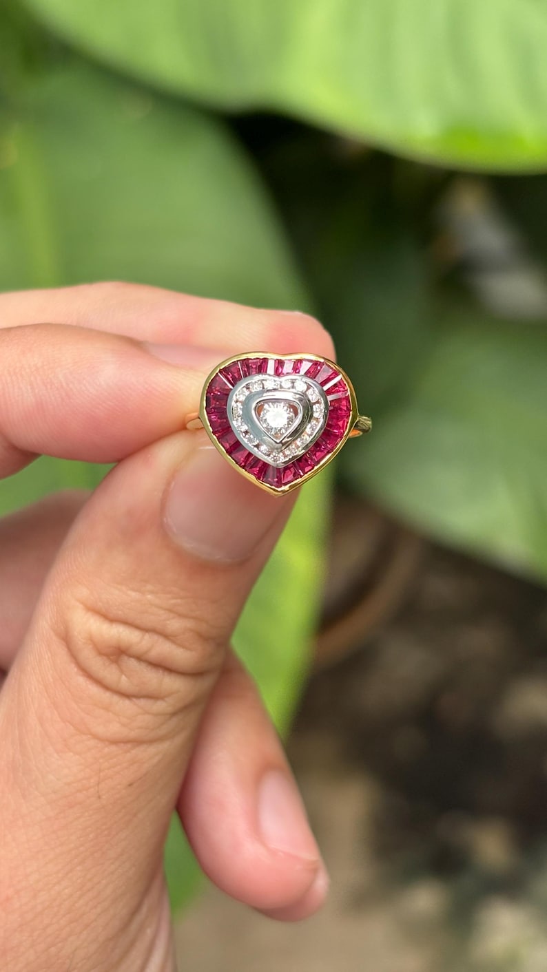 Pu&ograve; includere: Un anello a forma di cuore con una fascia d'oro. L'anello presenta un design centrale a forma di cuore con un diamante al centro, circondato da diamanti pi&ugrave; piccoli e pietre preziose rosse. L'anello &egrave; tenuto su uno sfondo verde sfocato.