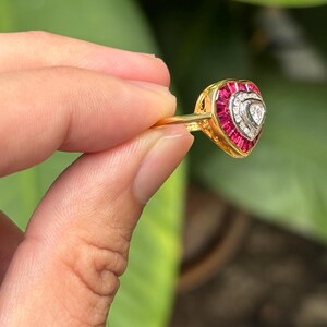 Pu&ograve; includere: Un anello d'oro con un design a forma di cuore. L'anello presenta un cuore centrale di pietre bianche, circondato da una fila di pietre rosse. L'anello &egrave; tenuto su uno sfondo verde.