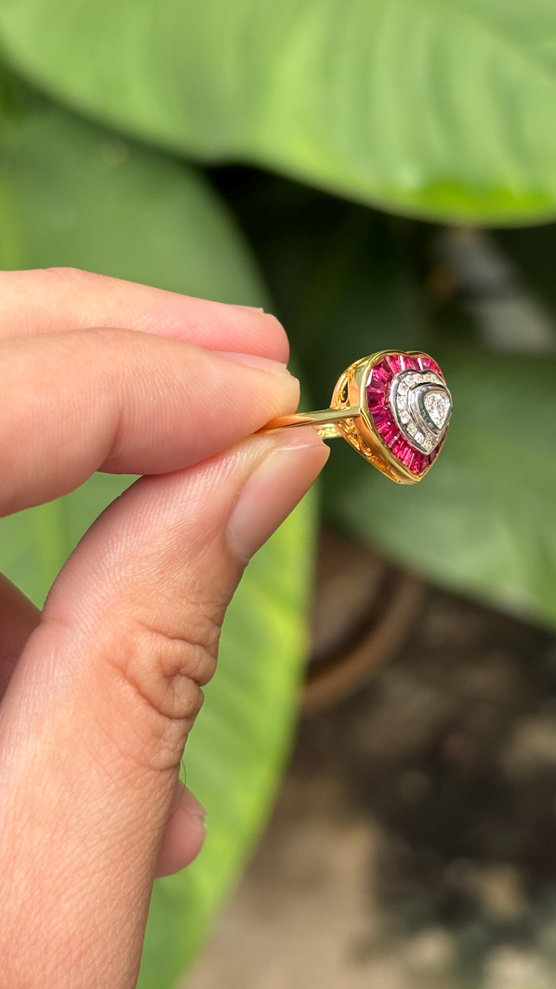 Pu&ograve; includere: Un anello d'oro con un design a forma di cuore. L'anello presenta un cuore centrale di pietre bianche, circondato da una fila di pietre rosse. L'anello &egrave; tenuto su uno sfondo verde.