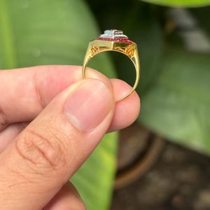 Pu&ograve; includere: Un anello d'oro con una parte superiore rettangolare con una pietra blu chiaro centrale, circondata da pietre rosse pi&ugrave; piccole. La fascia dell'anello &egrave; in oro lucido semplice. L'anello &egrave; tenuto contro uno sfondo di fogliame verde.