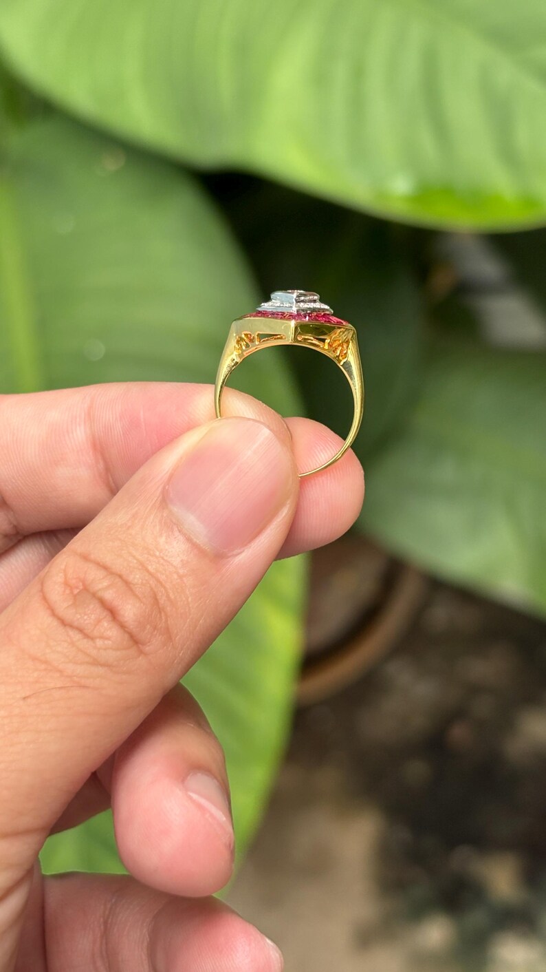 Pu&ograve; includere: Un anello d'oro con una parte superiore rettangolare con una pietra blu chiaro centrale, circondata da pietre rosse pi&ugrave; piccole. La fascia dell'anello &egrave; in oro lucido semplice. L'anello &egrave; tenuto contro uno sfondo di fogliame verde.