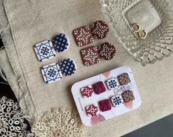 May include: A set of six handmade polymer clay earrings. The earrings are in two styles: a blue and white geometric pattern and a burgundy floral pattern. The earrings are displayed on a white card with the text "Handmade with love" printed on it.