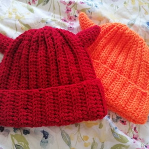 May include: Two crocheted hats with horns, one red and one orange. The hats are made with a textured stitch and have a pointed top.