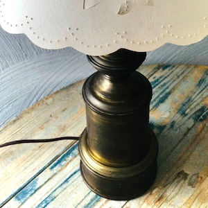 Puede incluir: Una lámpara de mesa de estilo vintage con una pantalla de color crema con un diseño de piña recortada. La lámpara tiene una base de color latón y un borde festoneado en la pantalla. La lámpara está sobre una superficie de madera.