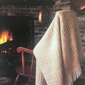 Op de afbeelding: Een witte geweven deken met franjes hangt over een houten schommelstoel voor een bakstenen open haard. De open haard heeft een brandend vuur en een koperen pot die erboven hangt.