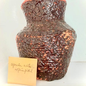 May include: A dark brown ceramic water jug with a rough, textured surface. The jug has a wide mouth and a narrow base. The jug is labelled "Apache water jug/Pine Pitch".
